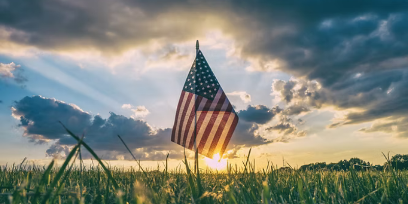 A lone American flag in a field of grass, set against the setting sun.